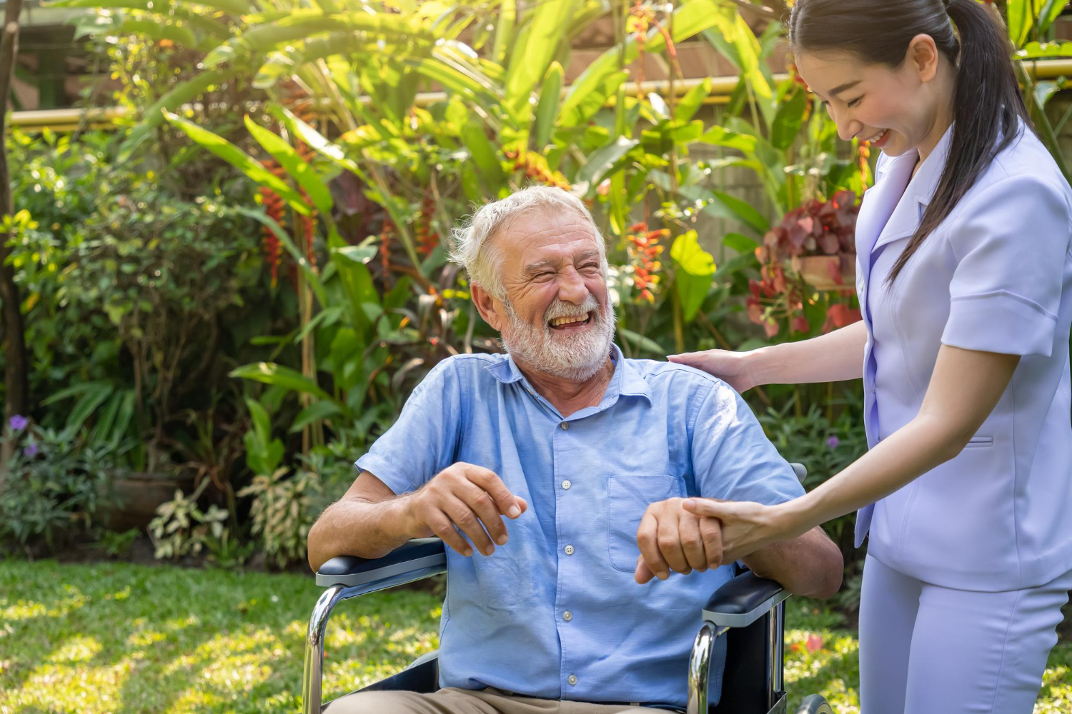 A cheerful nurse in uniform holding the hand of a smiling elderly man in a wheelchair out in a sunny, green garden.